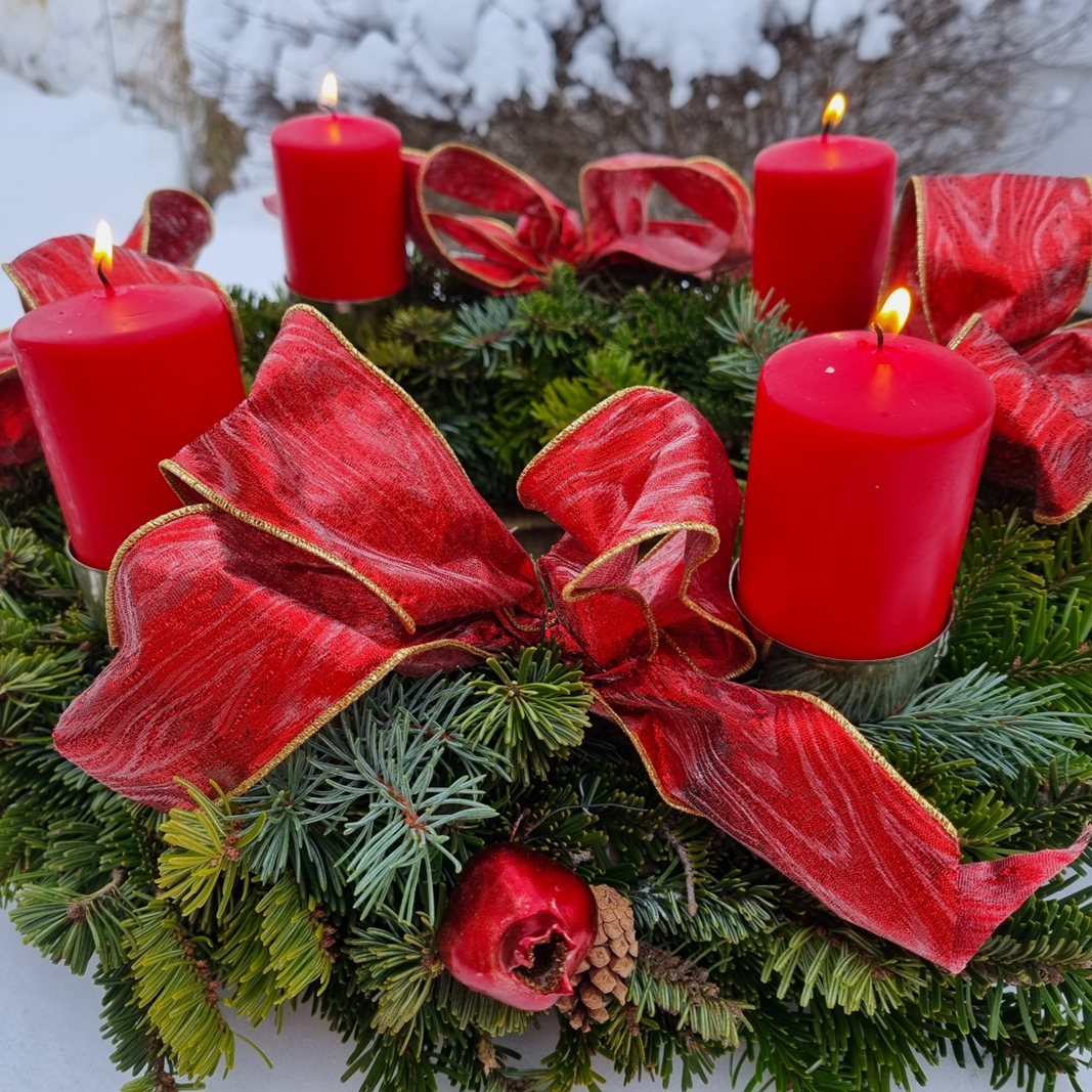 Ein Adventkranz mit roten Schleifen und brennenden roten Kerzen, steht auf einem Tisch im Schnee.